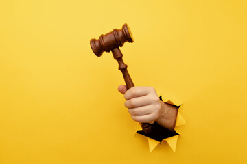 Hand holding a judge's gavel through torn yellow paper wall. Law and justice concept