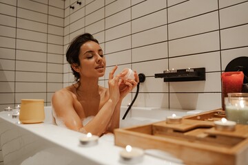 Young woman using mask on face, relaxing in the cosy bath full of foam and decorated with candles. Healthy treatment and enjoyment.