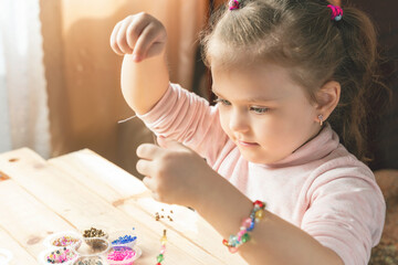 A little girl is engaged in needlework, making jewelry with her own hands, stringing multi-colored...