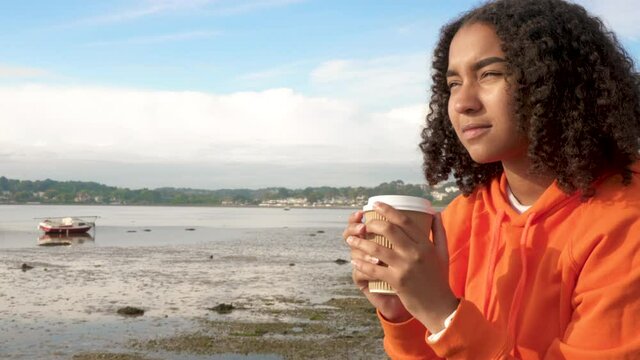 Beautiful mixed race African American girl teenager young woman wearing orange hoodie, drinking takeout coffee by the sea looking sad or thoughtful then happy smiling