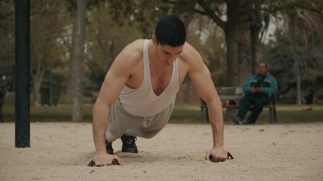 hombre haciendo flexiones en el parque, hombre de pelo negro haciendo deporte al aire libre, hombre musculoso en camiseta de tirantes  blanca haciendo ejercicio