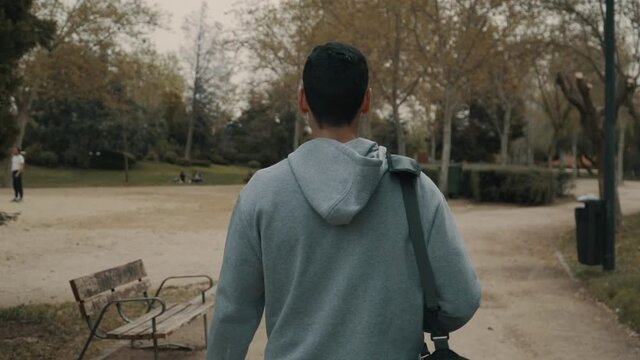 Hombre de pelo negro caminando con su bolsa de deporte, hombre con sudadera gris y ch&aacute;ndal caminando, deportista caminando por el parque, c&aacute;mara siguiendo al deportista desde atr&aacute;s