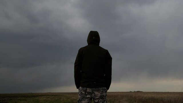 Rear view of male person wearing hooded jacket against dark moody dramatic clouds at sky
