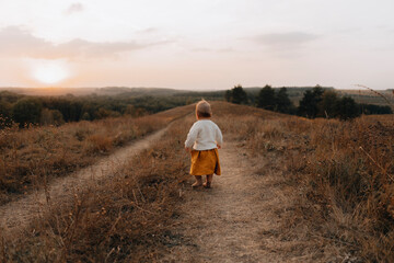 Naklejka na ściany i meble little girl walks along path at the top of the hill. baby without shoes walks on top of a hill in the forest at a beautiful sunset. a lonely child walks along a path , view from the back. copy space