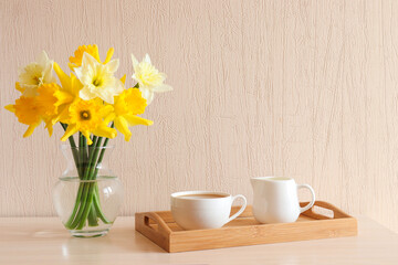 Cozy home interior. Delicate yellow daffodils in glass vase and cup of coffee with milk on a wooden tray on the table. Copy space. Springtime concept