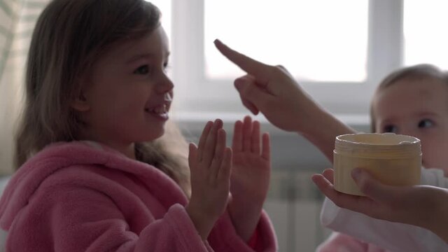 Authentic cute young woman applying face mask of baby girl. mom with 2 children daughters different ages in bathrobes after bath on white bed oposite window. Spa, motherhood, childhood, family concept