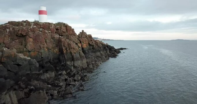 4k aerial fly past over sea past Aberdour Lighthouse in Fife, Scotland showing red and white lighthouse, rocky cliffs and Firth of Forth estuary. Early morning light.