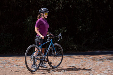 Chica con su bicicleta de pista, usando casco y lentes de sol, junto a la carretera en medio del...
