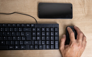A dark man's hand on a computer mouse with a black keyboard on the side and a cell phone in front of a screen messenger used for work in the home office. Detail of the numeric keys.
