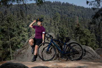 Fototapeta premium Chica ciclista recargado de una piedra con su bicicleta al lado, con un paisaje al fondo. Deporte al aire libre