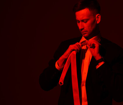 Portrait of young focused businessman in official wear, suit tying his tie looking at it over black background with free copy space. Red light filter, gel