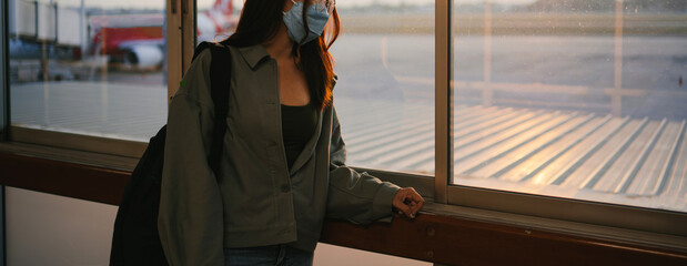 Traveler business woman wearing face mask looking to airplane, standing with backpack in departure terminal in airport. Female passenger flight traveling by plane during covid19 virus pandemic.