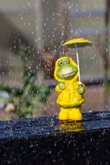 figure of a frog with yellow rain jacket stands in spring in rainy weather and is protected by a yellow rain umbrella