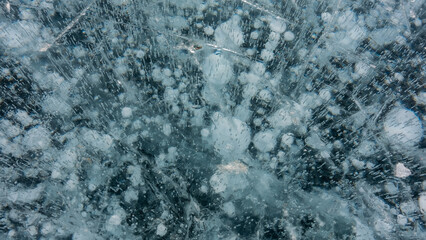 Transparent ice of Lake Baikal, close-up. Cracks are visible on the smooth turquoise surface, in the depth there are columns of white bubbles of frozen methane gas. Full frame