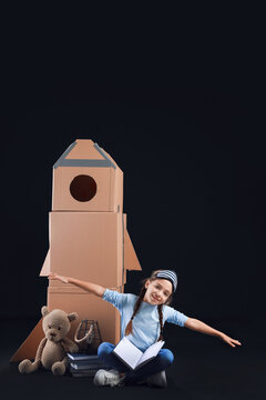 Cute little girl with cardboard rocket, books and toy on dark background