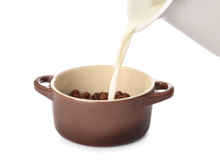 Pouring of milk from jug into cooking pot with chocolate corn balls on white background