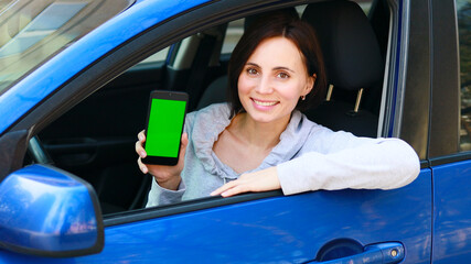 European woman with short hair in grey clothes sitting in the blue car, smiling and showing a mobile phone with green screen