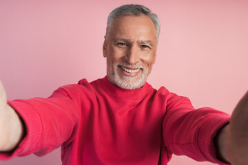 Close-up shot of a senior man's hands holding his phone, taking a selfie, on a pink background. Smiling man isolated on pink background.