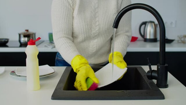 Close-up of female hands in yellow protective rubber gloves washing dishes after meal using dish detergent and sponge in domestic kitchen during housework.