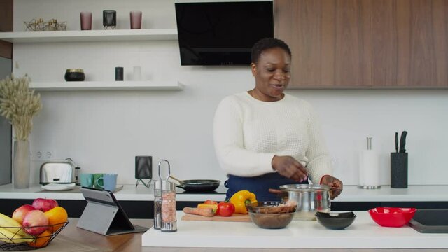 Positive Attractive Black Woman In Apron Having Video Call Online Using Digital Tablet, Communicating And Preparing Meal, Expressing Positivity And Happiness While Enjoying Cooking In Domestic Kitchen