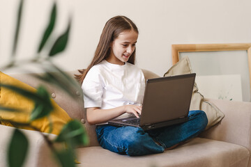 A smiling dark-haired teenage girl sitting at home on the couch,using a laptop,surfing social...