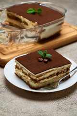 Traditional Italian Tiramisu dessert in glass baking dish and portion on grey concrete background