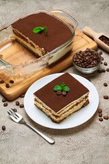 Traditional Italian Tiramisu dessert in glass baking dish and portion on grey concrete background