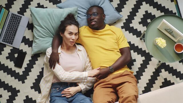 Directly above shot of Afro-American man embracing young mixed race wife while lying together on floor in living room and looking at camera