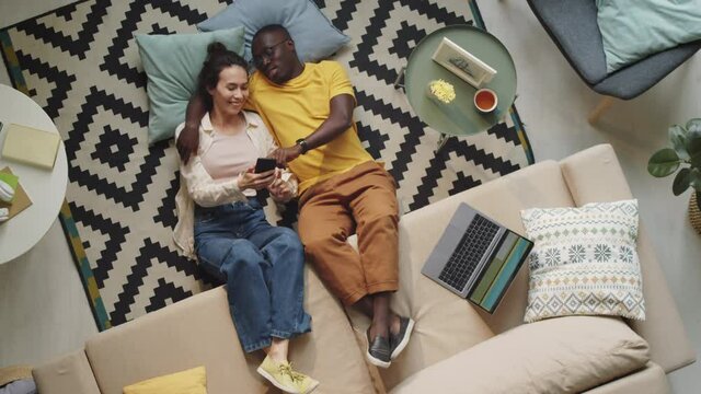 Top down shot of multiethnic family couple lying on floor in living room with legs on sofa, embracing, using smartphone and speaking while spending day together at home