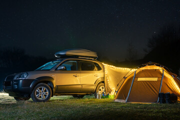 Fototapeta na wymiar Night landscape with illuminated tent and 4wd car, lights and stars in the background. Spring camping. Tourism and vacation. Roof rack, car luggage box