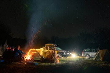 Group of travelers sitting near bonfire, cars and camp tent under beautiful night sky. View of blue starry sky over meadow with travelers near campfire.