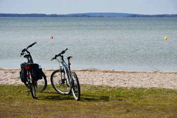 Fototapeta na wymiar Close up on two bikes with bags standing next to a sandy coast of a river or lake with some red and yellow buoys visible in the distance seen on a sunny spring day on a Polish countryside 
