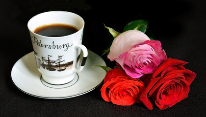 A white cup with black coffee is on a saucer. Nearby lies a bouquet of three beautiful multi-colored rose buds. On a dark brown background.
