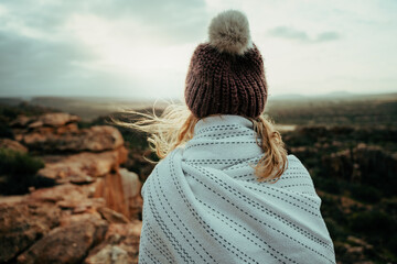 Caucasian female camping on top of mountain wrapped in blanket wearing beanie in the cold weather 