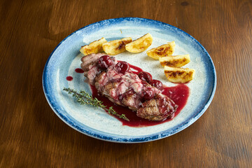 Delicious baked confit duck breast with berry sauce and potatoes, served on a blue plate. Wood background