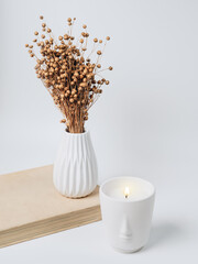 A beautiful arrangement of dried flowers in a stylish ceramic vase and a candle in a white candlestick. A bouquet of dry flax on a light background. Minimal modern interior decoration.