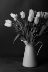 Tulips in a jug. Classic still life with a bouquet of tulip flowers in a vintage white jug on a dark background and an old wooden table. The style is black and white.