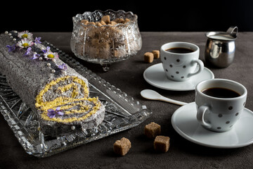 Traditional Czech sweet roll with poppy seed filling and edible flowers served with coffee decorated with brown cane sugar cubes on a dark grey stone background