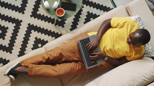 Overhead shot of Afro-American man lying on sofa and typing on laptop while working online at home