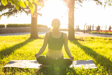 Cute young caucasian pregnant woman is meditating while sitting on a rug on the lawn on a sunny summer evening. Concept of pacification and energy boost