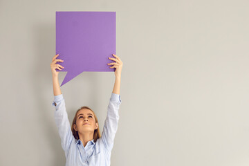 Have your say. Happy young woman or teen girl holding up one clean violet square shaped free copyspace speech bubble on gray background. Inspirational quote, message, expressing opinion mockup card