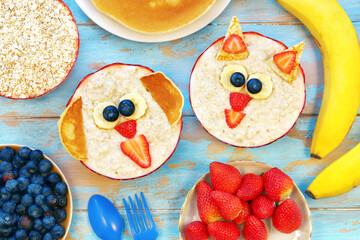 Obraz premium Creative idea for kids breakfast. Cute oatmeal cat and dog with berries on blue wooden table, top view. Healthy vegetarian eating for children