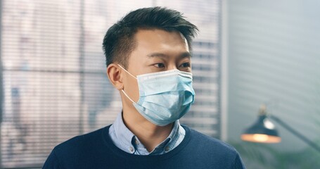 Close up portrait of handsome young Asian male employee in medical mask looking at camera in cabinet indoor. Businessman at office in quarantine. Business, work, working during covid-19 pandemic