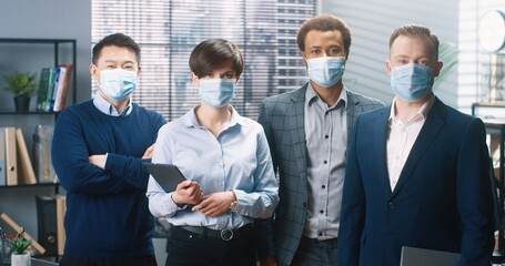 Portrait of positive young joyful mixed-race workers standing at office in international company, looking at camera and smiling. Men and woman in medical masks in cabinet, business concept