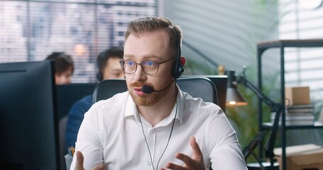 Close up portrait of upset young Caucasian male worker speaking in headset on video call on computer sitting at table at office with sad tired face. Handsome man employee talk on online call