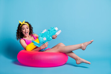 Full body profile side of young excited girl happy smile play game water gun sit on float circle isolated over blue color background