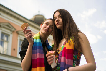 A young lesbian couple embraces their love. They are walking the streets and eating ice cream covered together with a rainbow colors scarf.