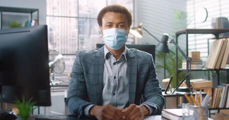 Fototapeta na wymiar Portrait of African American handsome young businessman in medical mask sitting at table at workplace, typing on computer browsing online. Professional working in cabinet. Job concept