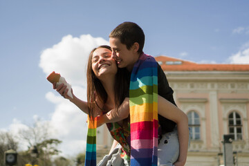 A young lesbian couple embraces their love. They are walking the streets and eating ice cream covered together with a rainbow colors scarf. Piggyback ride.