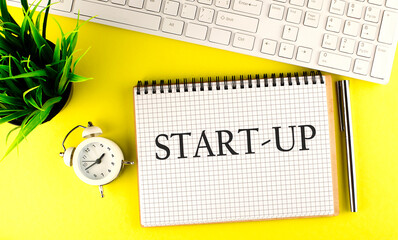 START UP text on notebook with keyboard , pen and alarm clock on the yellow background